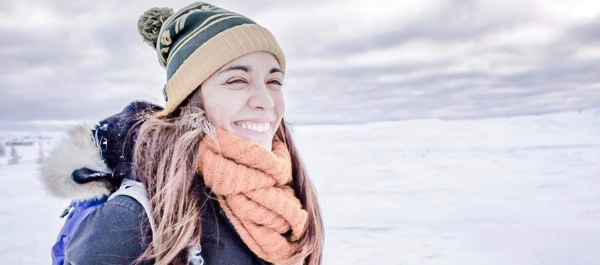 Woman in winter jacket and hat stands outside in a snowy northern environment smiling.